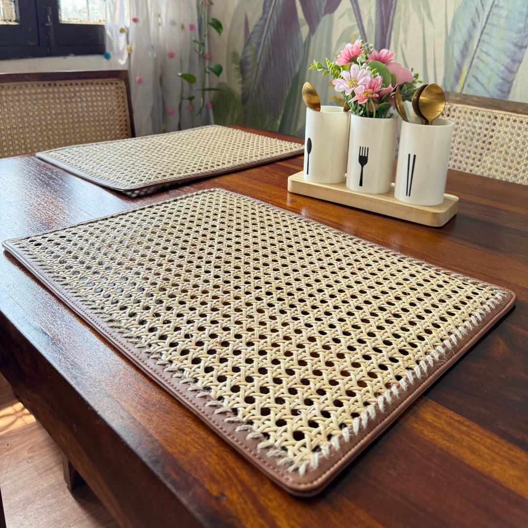 Rectangular rattan table mats placed on a wooden dining table, with one mat partially covering the other. The mats have a beige color with a woven rattan pattern and dark brown vegan leather edging.