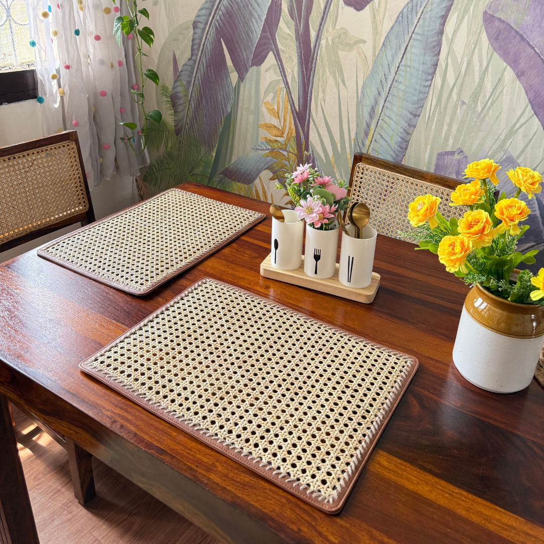 Rectangular rattan table mats placed on a wooden dining table with floral wallpaper in the background. The mats are beige with a woven pattern and have a rectangular shape.