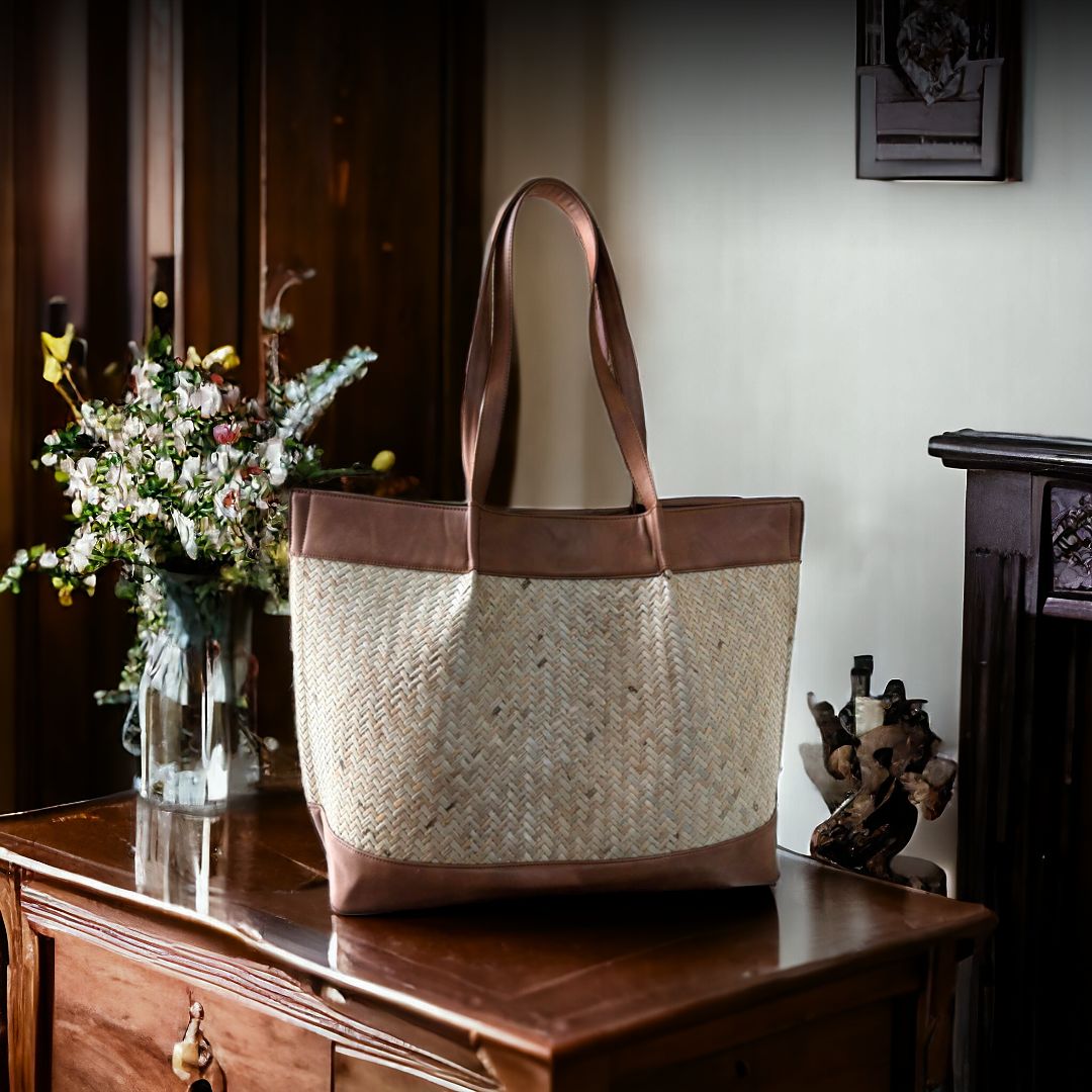 A beige tote bag with wicker design and brown vegan leather accents, displayed on a wooden table alongside a vase of flowers.