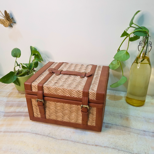 A brown wicker jewellery box with a chevron pattern, placed on a marble surface, with a plant and a glass bottle in the background.