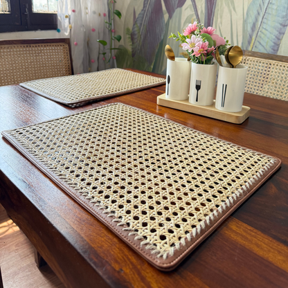 Rectangular rattan table mats placed on a wooden dining table, with one mat partially covering the other. The mats have a beige color with a woven rattan pattern and dark brown vegan leather edging.