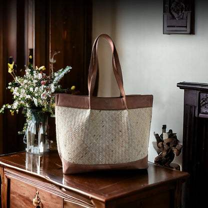 A beige tote bag with wicker design and brown vegan leather accents, displayed on a wooden table alongside a vase of flowers.
